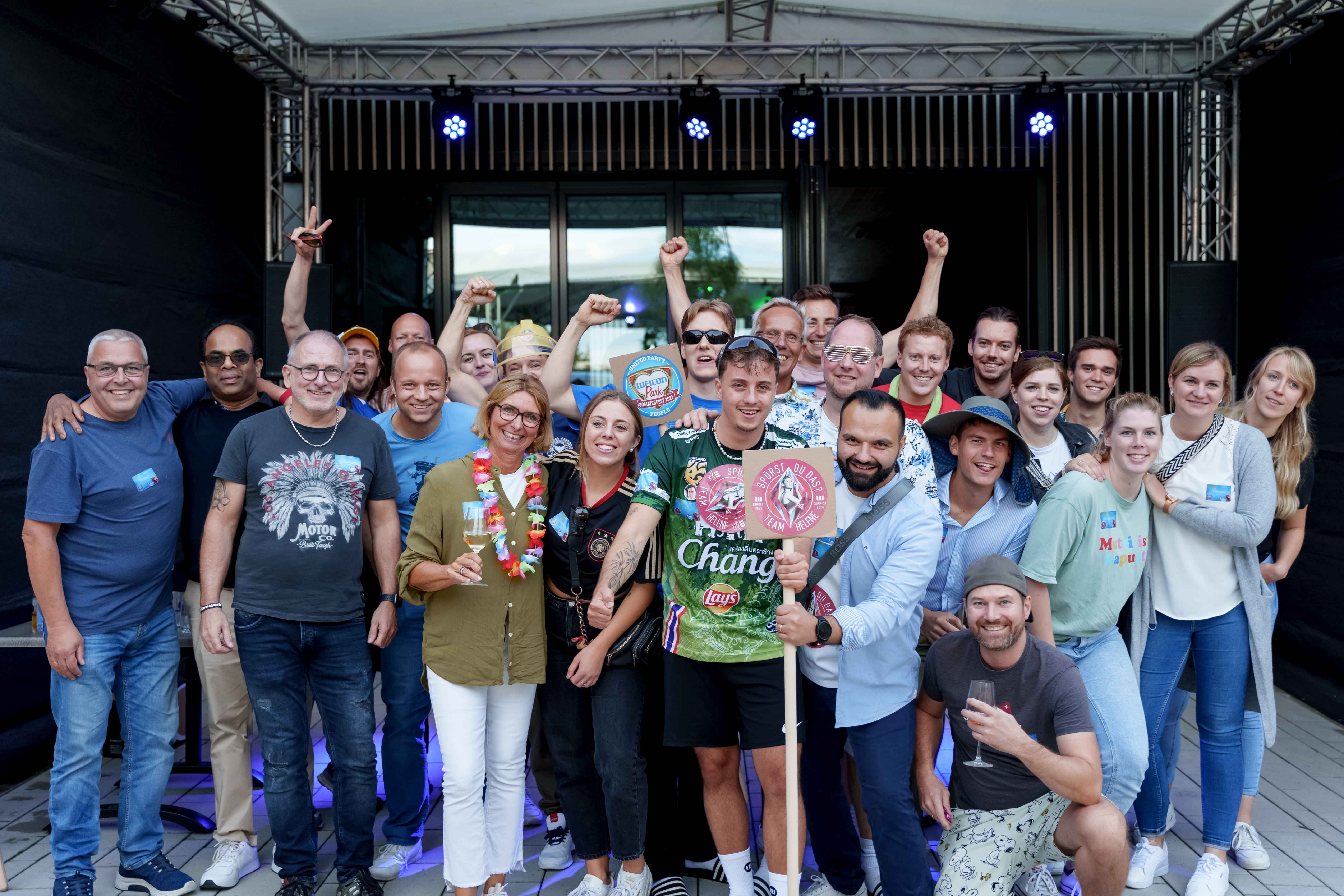 eine Gruppe von ca. 20 Menschen steht draußen vor einer Bühne und lacht in die Kamera