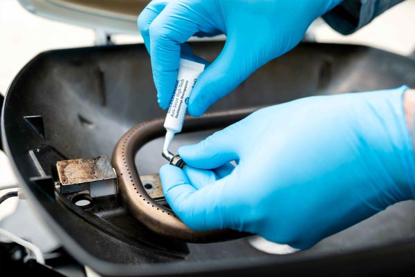 Application d'une pâte anti-grippante Anti-Seize WEICON sur un petit composant métallique au-dessus d'un brûleur à gaz par une personne portant des gants bleus.