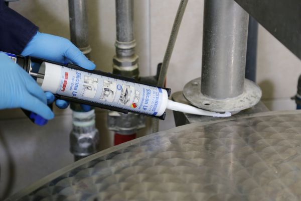 Application d'un mastic silicone WEICON avec un pistolet extrudeur par un opérateur en gants bleus sur une structure métallique industrielle.