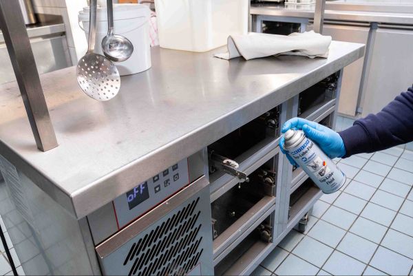 Application d'un lubrifiant WEICON en spray sur les mécanismes d'un meuble de cuisine en acier inoxydable par un opérateur en gants bleus.