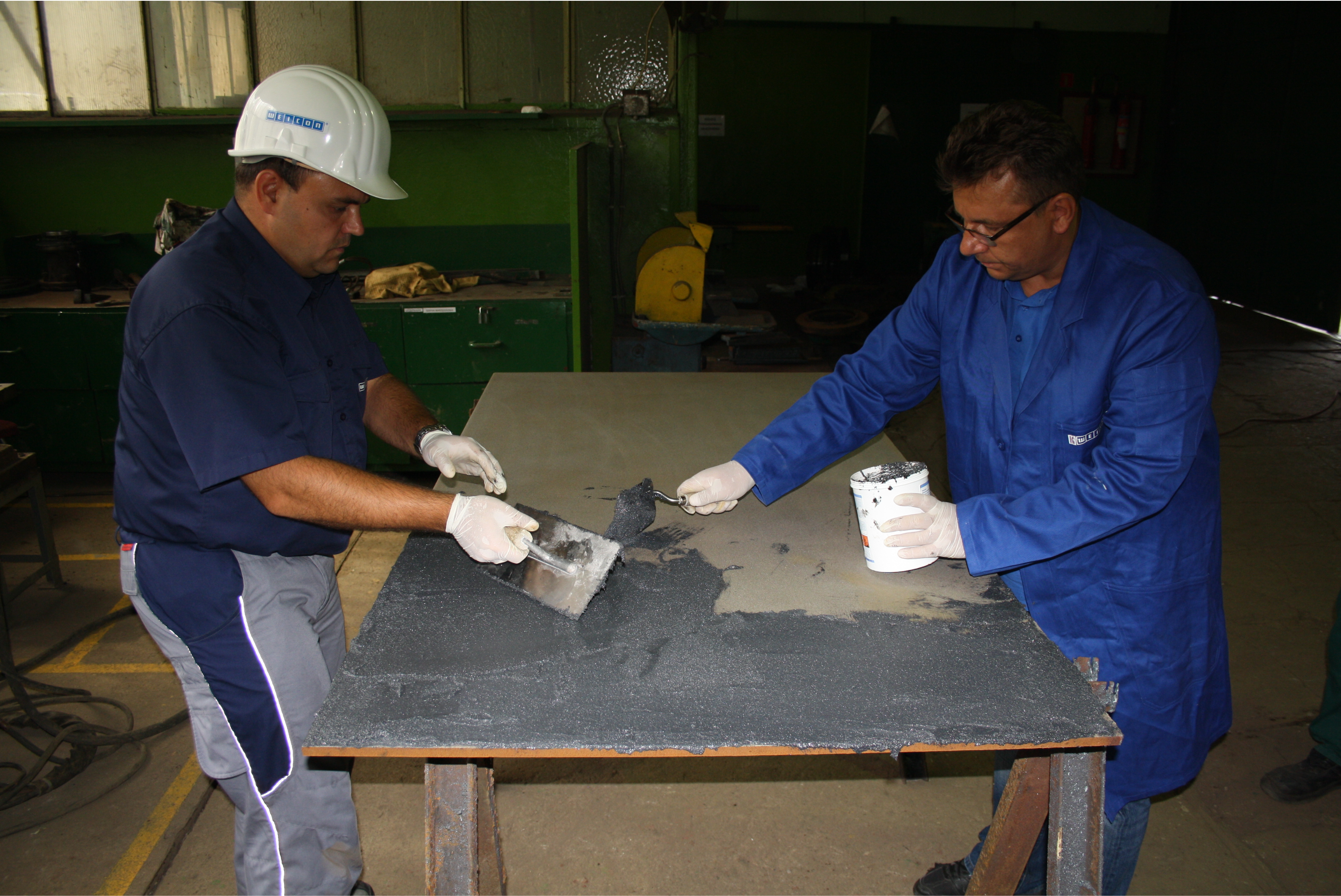 Due lavoratori in un'officina, entrambi con guanti protettivi, applicano e spalmano un composto denso e scuro su una grande lastra di metallo posta su un tavolo da lavoro;