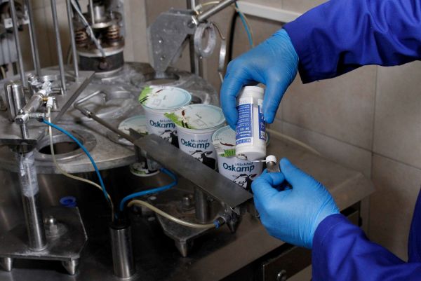 Application d'un produit de maintenance WEICON sur une pièce mécanique au-dessus d'une ligne d'emballage de pot de yaourt par un opérateur en gants bleus.