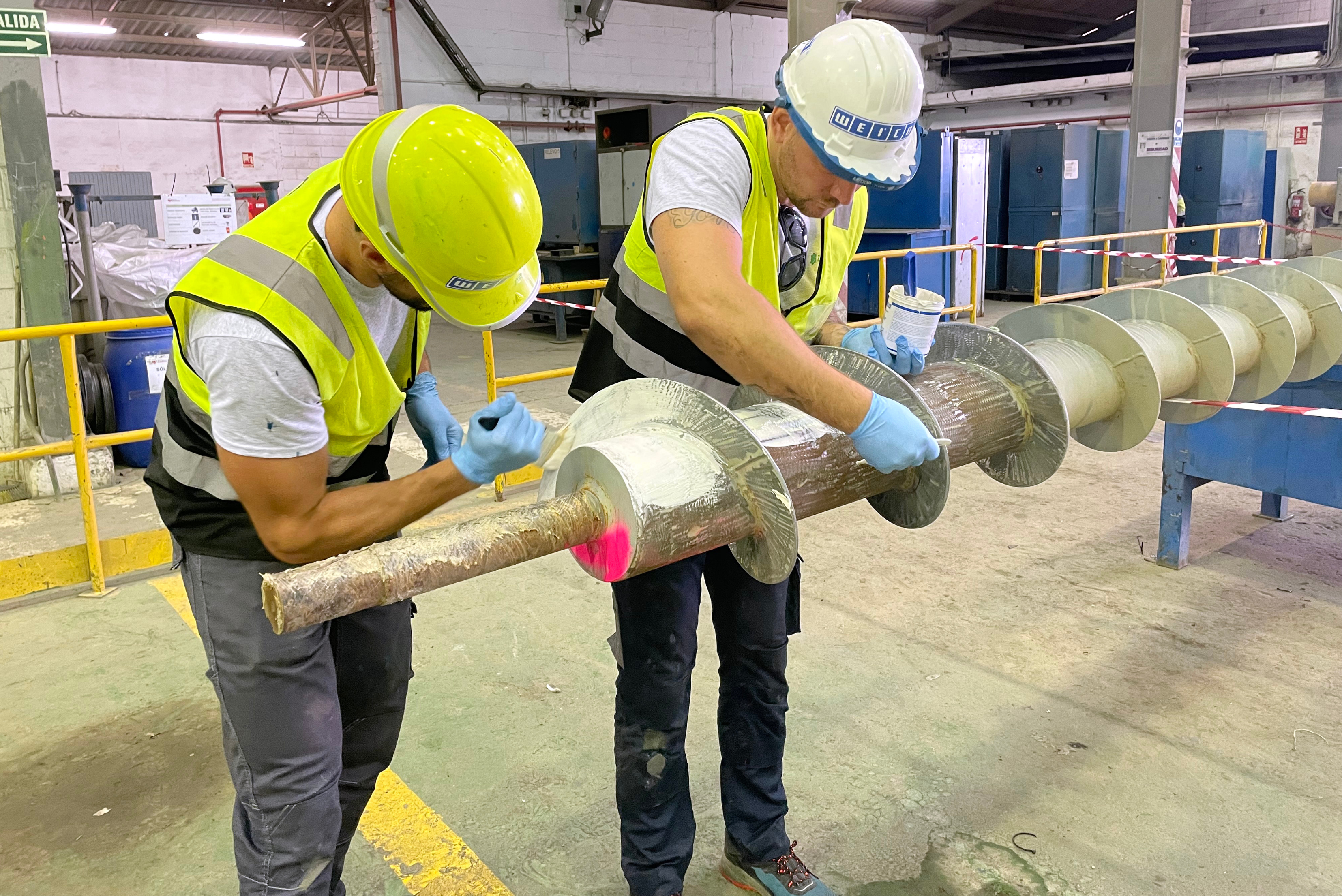 Due lavoratori con elmetti di sicurezza, giubbotti ad alta visibilità e guanti protettivi applicano un rivestimento protettivo su una grande coclea industriale o rotore all'interno di un'officina.
