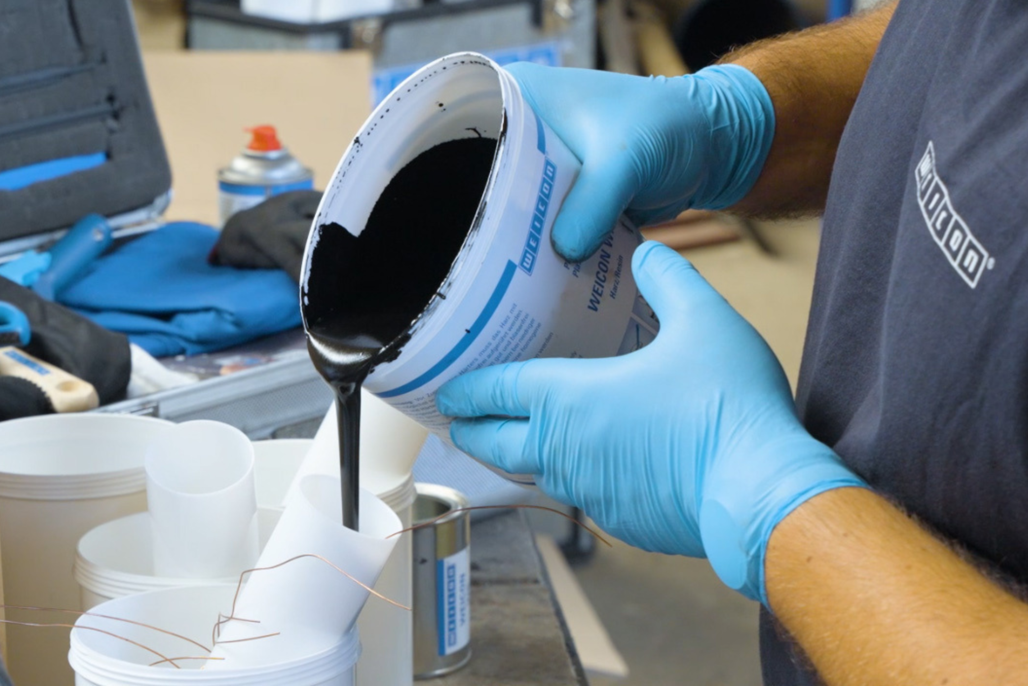 Una persona che indossa guanti protettivi azzurri versa un liquido denso e scuro da un secchio bianco con dettagli blu in un contenitore più piccolo posto su un banco da lavoro.