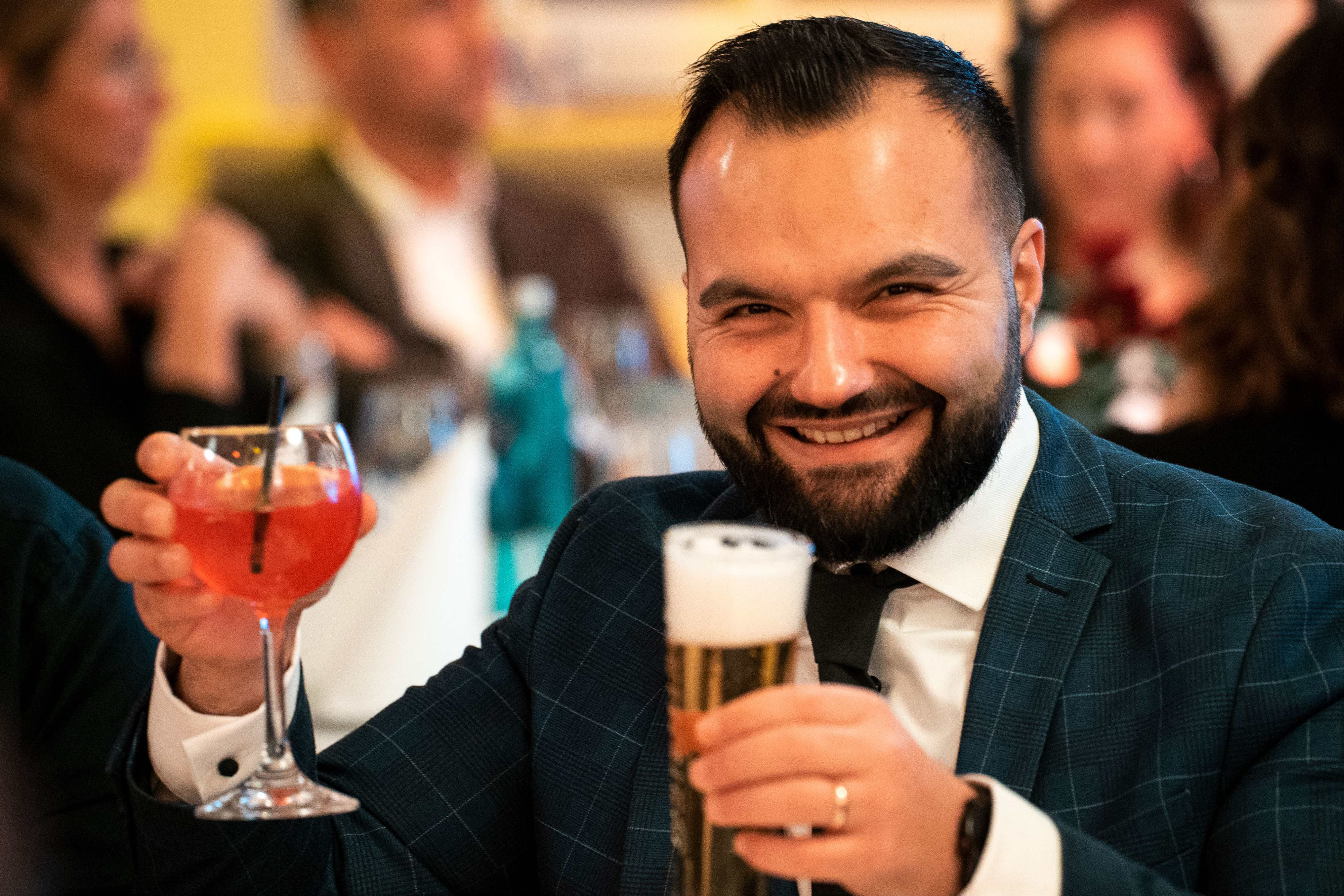 Ein Mann in Anzug lacht auf einer Veranstaltung in die Kamera und hält in der einen Hand ein Bier und in der anderen einen Aperol