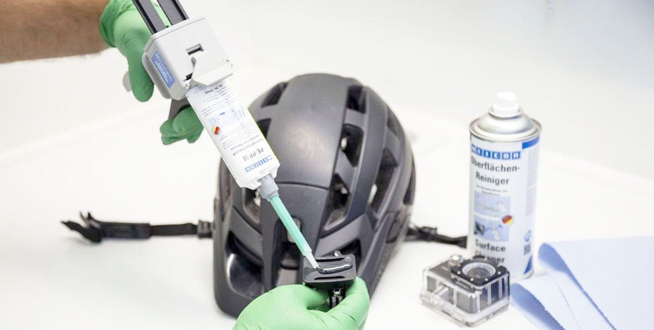 Der Plastikkleber Weicon PE-PP wird auf die Halterung einer Helmkamera gegeben. Dahinter liegen der Helm und die Kamera. Daneben steht ein Weicon Oberflächenreiniger.