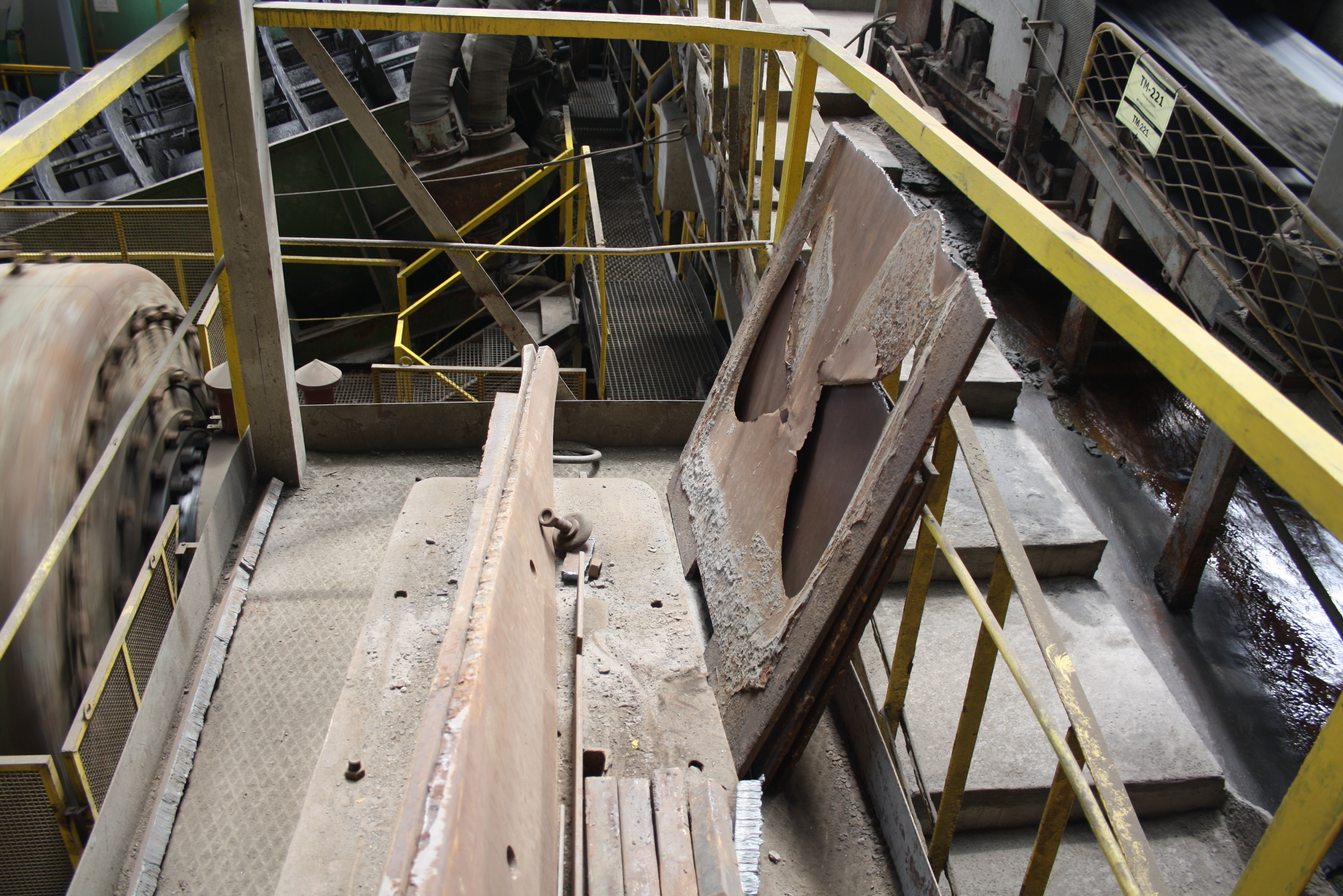 Una vista dall'alto mostra una passerella industriale con ringhiere gialle, su cui sono appoggiate lamiere e componenti metallici arrugginiti accanto a macchinari in funzione.