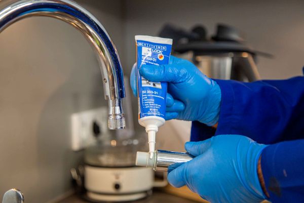 Application d'un produit d'étanchéité fileté WEICON Lock sur un raccord de tuyauterie par un opérateur en gants bleus près d'un robinet.