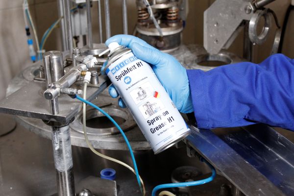 Application d'une graisse en spray WEICON Sprühfett H1 sur un mécanisme industriel par un opérateur en gants bleus.