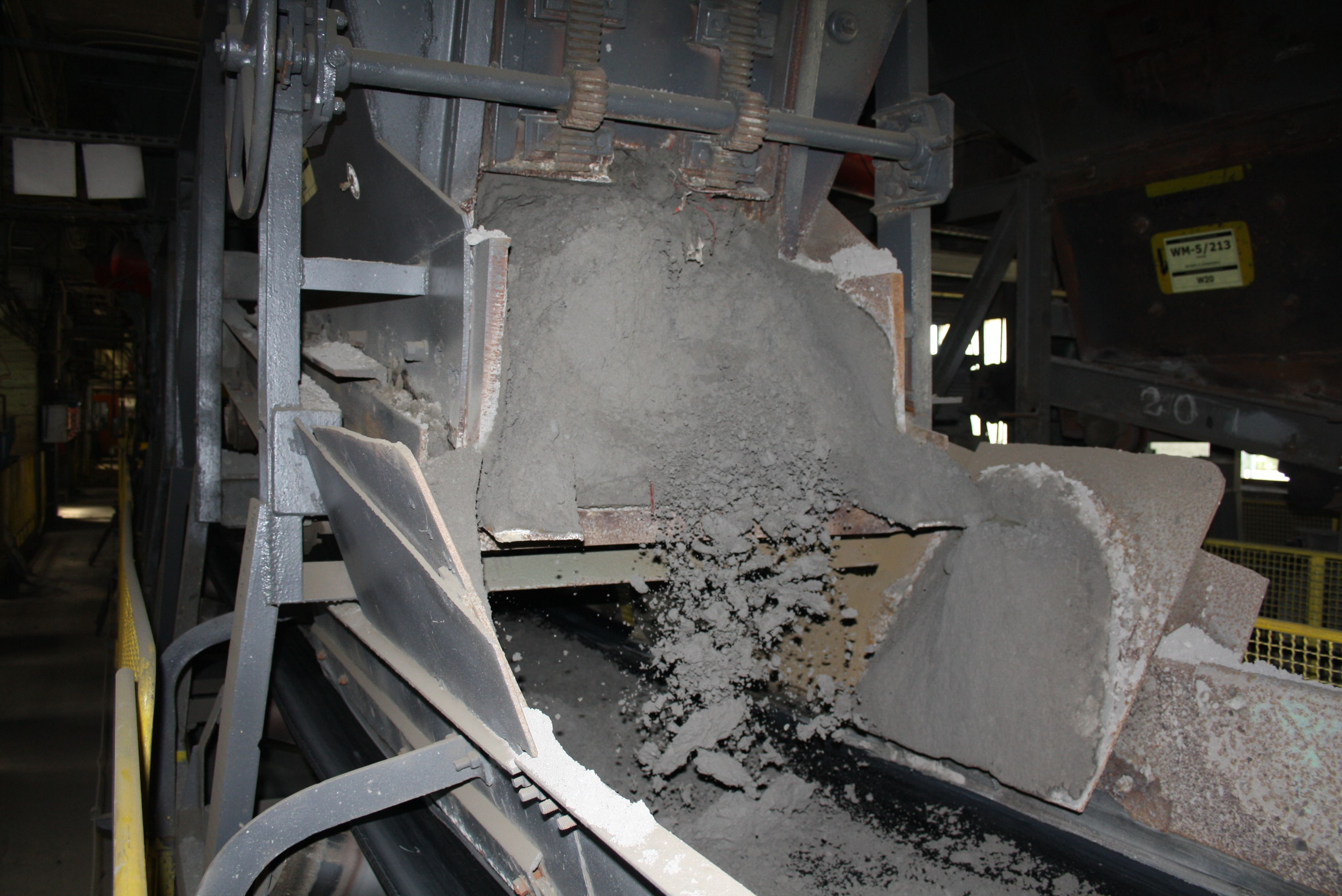 Un nastro trasportatore industriale in un impianto movimenta materiale grigio e polveroso che cade da una tramoggia su un nastro trasportatore sottostante.
