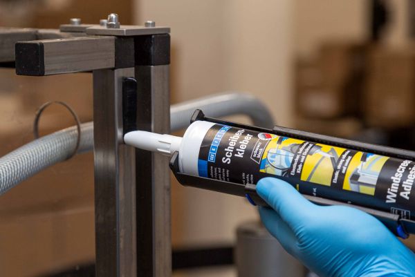 Application d'une colle à pare-brise WEICON avec un pistolet extrudeur par un opérateur en gants bleus sur une structure métallique.