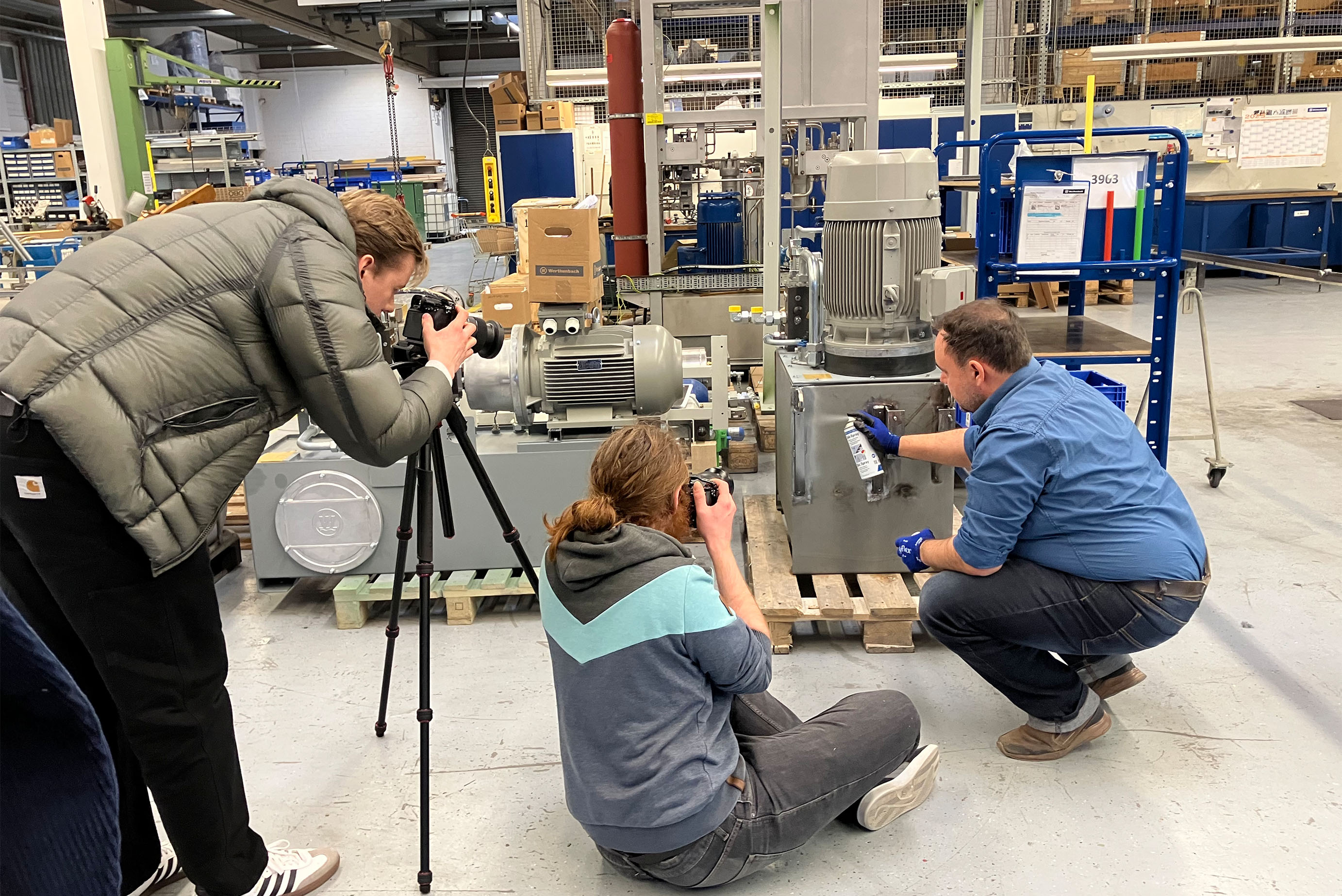 Drei Männer sind in einer Halle, steht an einem Stativ und fotografiert, einer sitzt auf dem Boden und fotografiert und der Dritte bückt sich und sprüht etwas auf eine Anlage