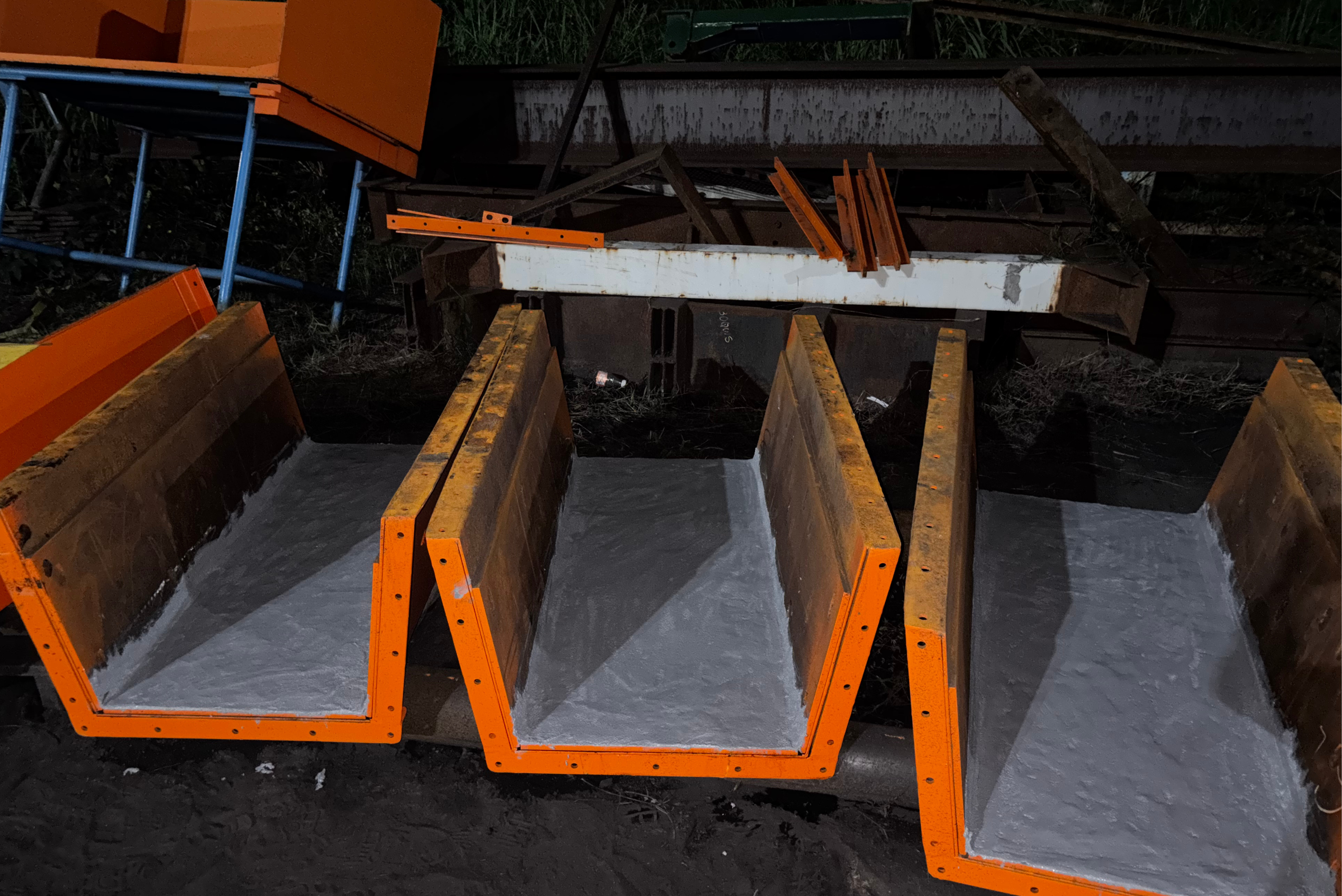 Tre canali metallici a forma di U, con bordi arancioni brillanti e interni rivestiti di uno strato protettivo grigio chiaro, sono disposti uno accanto all'altro in un'area di lavoro all'aperto.