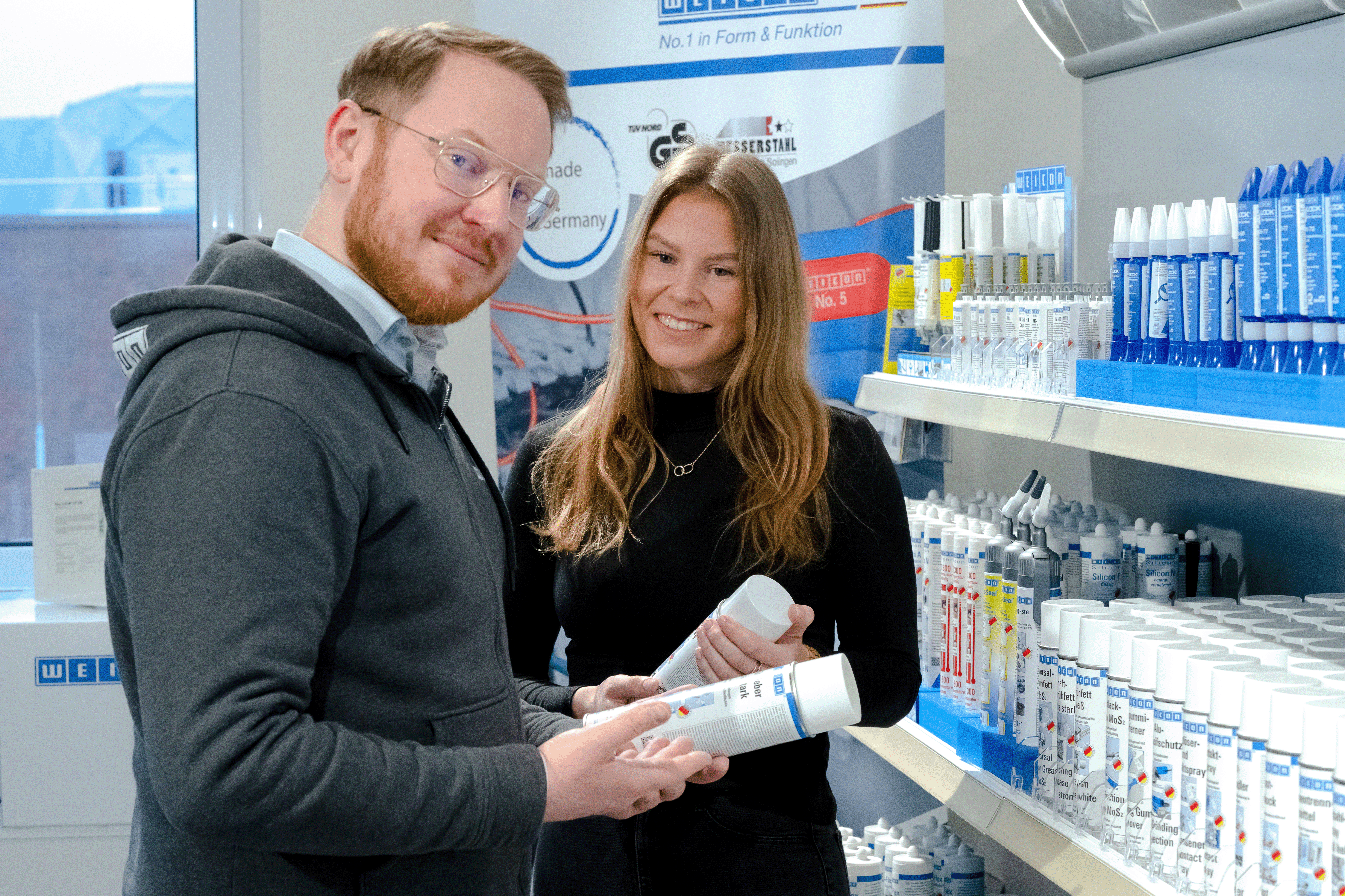 Ein Mann hält eine Spraydose in der Hand und lacht und die Kamera, eine Frau steht daneben hat eine Spraydose in der Hand und lächelt auch in die Kamera, daneben ist ein Regal mit Weicon Produkten