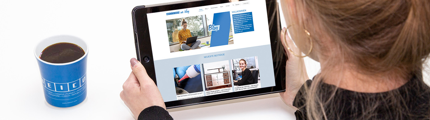 online Hinterkopf einer Frau, sie hält ein Tablet in der Hand und schaut sich den Weicon-Blog an, neben ihr steht eine blaue Weicon-Tasse mit Kaffee