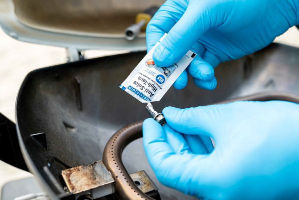 Application de pâte Anti-Seize High-Tech WEICON depuis un sachet sur une vis filetée au-dessus d'un système de brûleur à gaz par un technicien en gants bleus.
