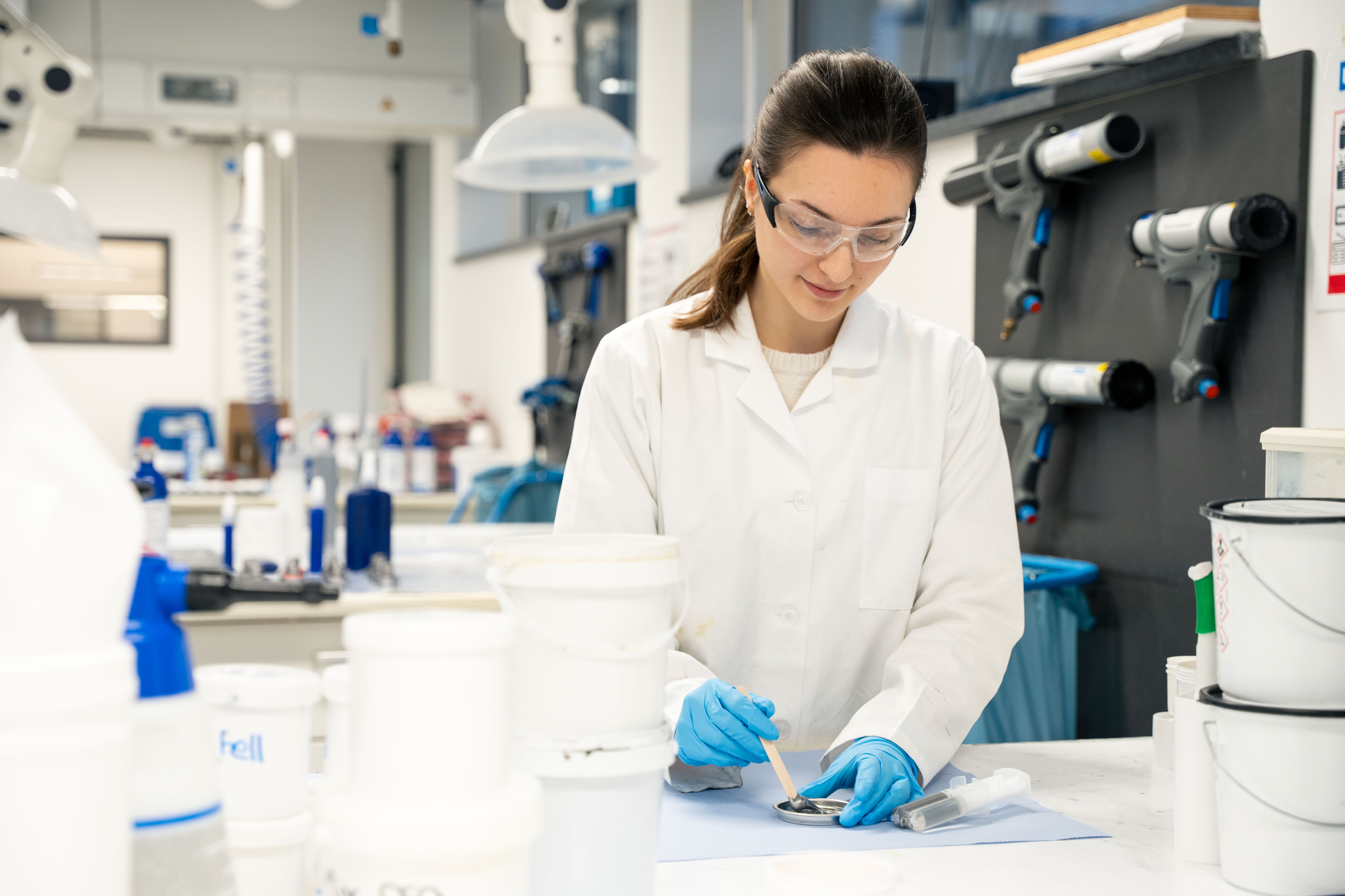 Eine Frau steht im Labot mit weißem Kittel, blauen Handschuhen und Brille und vermengt einen Klebstoff