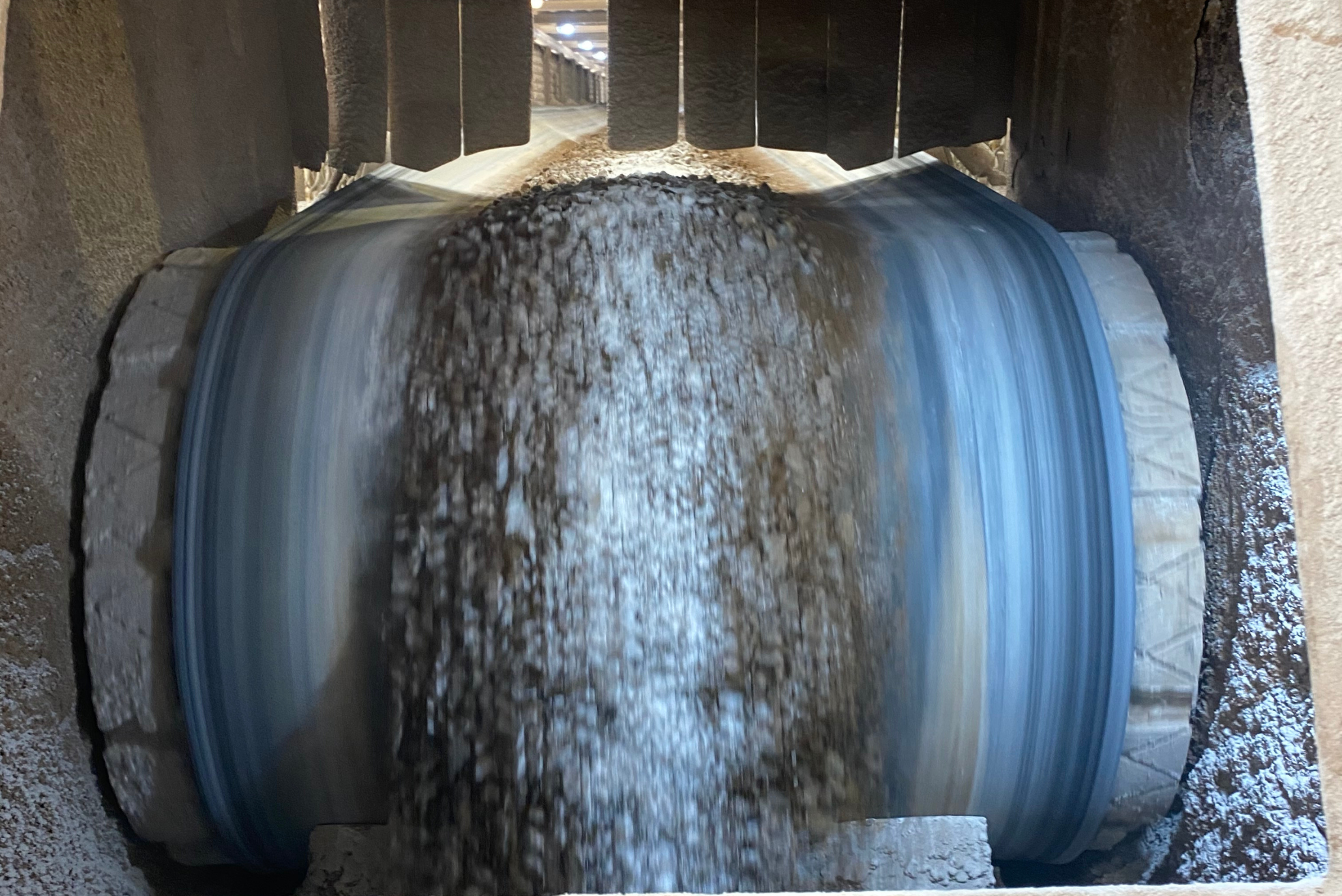 Un nastro trasportatore industriale in movimento trasporta del materiale sfuso pietroso all'interno di un impianto.