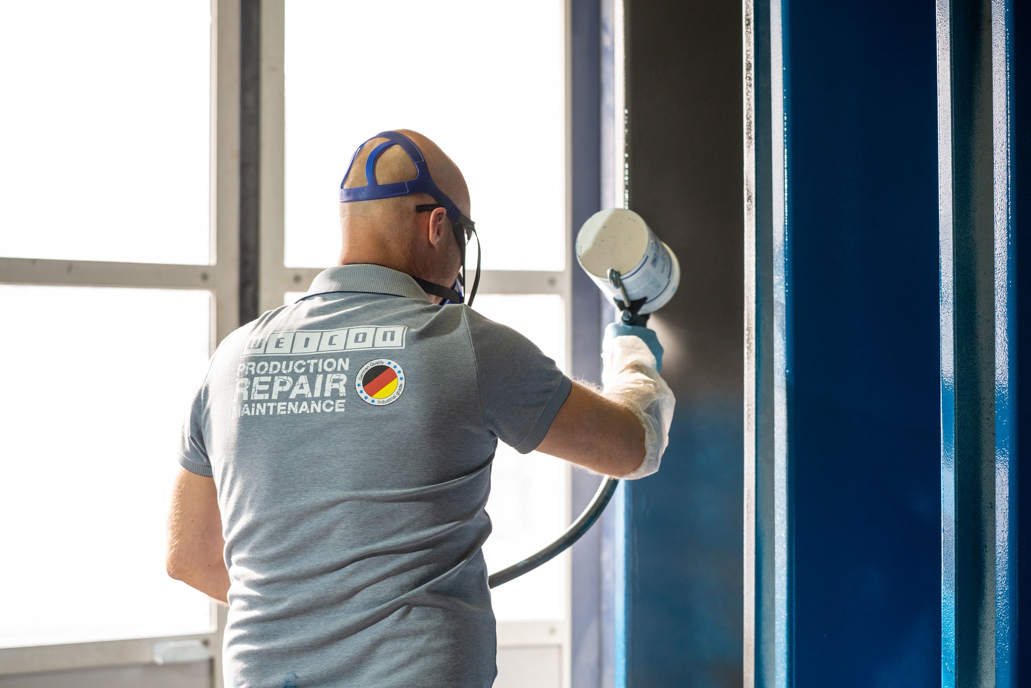Un tecnico di spalle, che indossa una maglietta grigia con la scritta WEICON e dispositivi di protezione individuale come una maschera e guanti, utilizza un'attrezzatura a spruzzo per applicare un rivestimento su componenti industriali metallici.