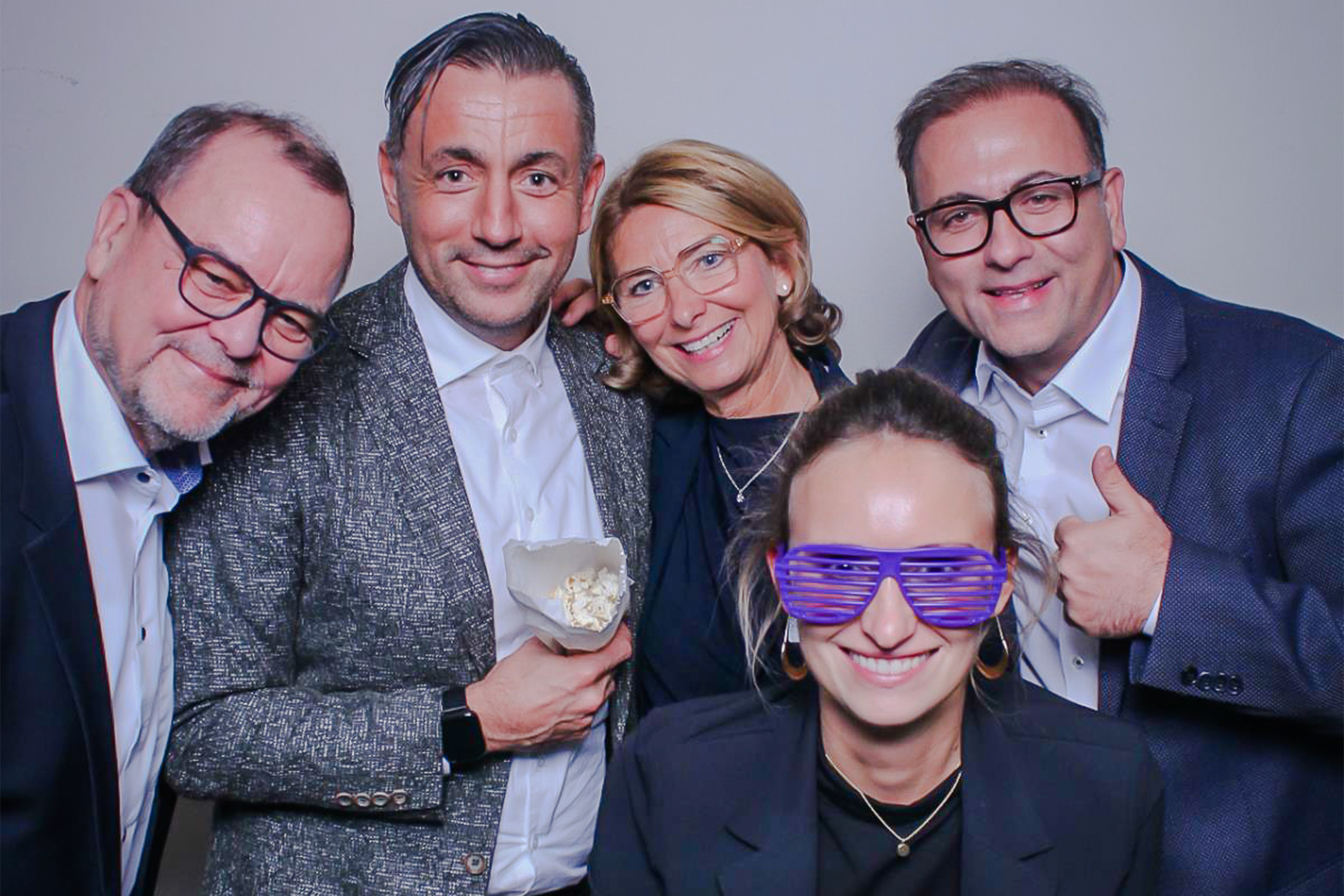 Fünf schick gekleidete Personen stehen nebeneinander und lachen in die Kamera, eine trägt eine lilane Sonnebrille, einer hat ein Weinglas in der Hand