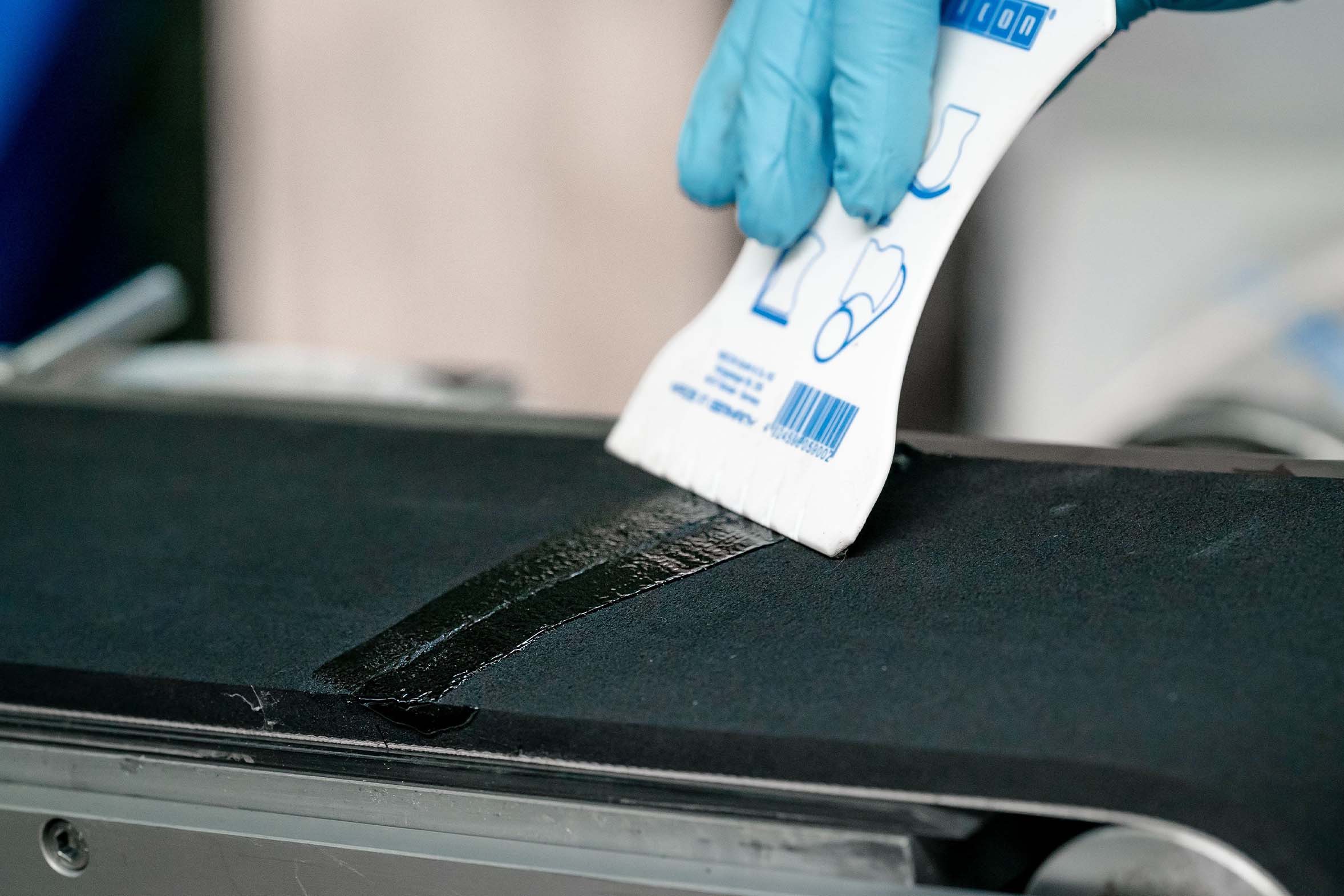 schwarze Vergussmase wird mit einem weißes Spatel auf defektem Förderband glatt gestrichen