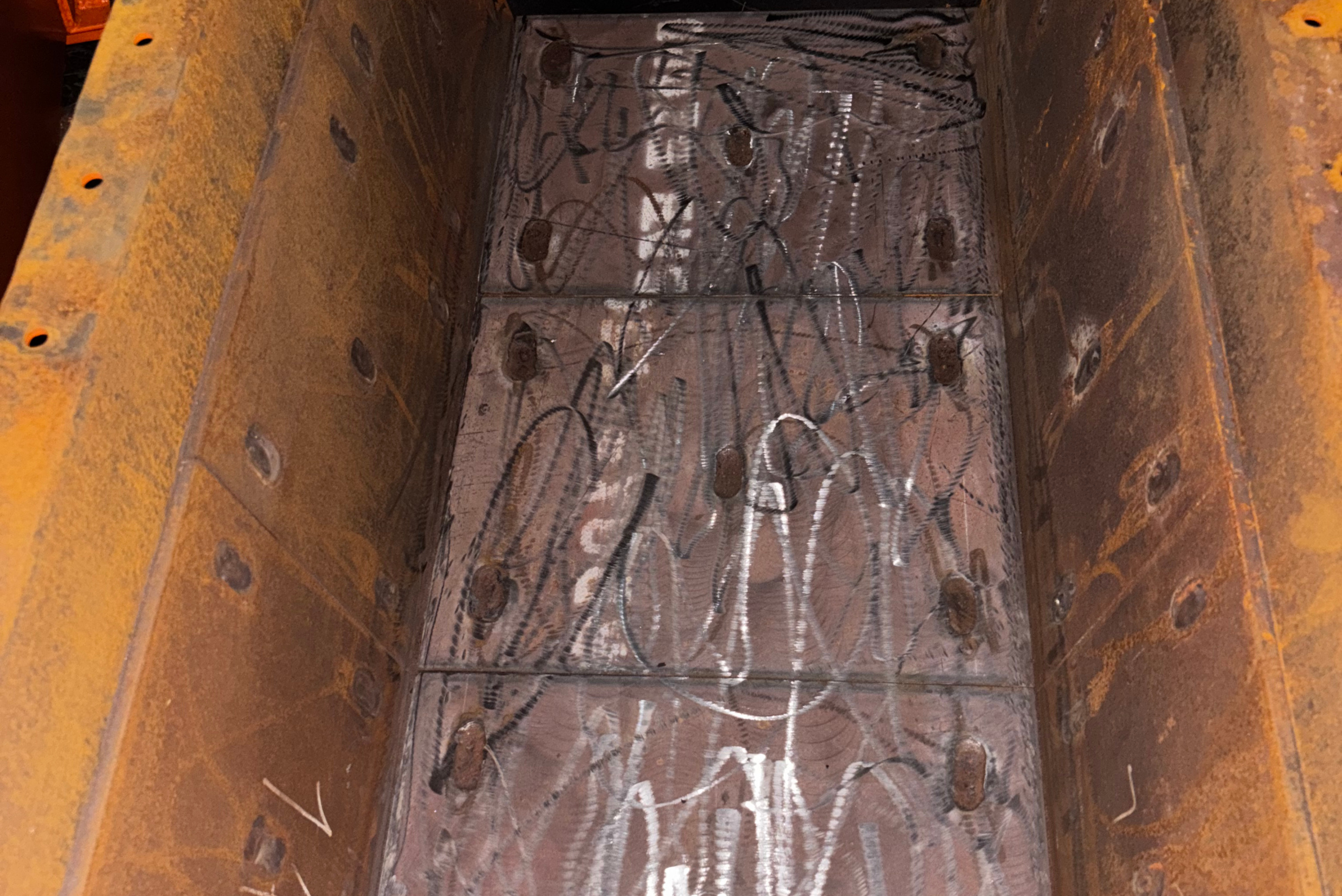 Un'inquadratura dall'alto mostra l'interno di un cassone metallico arrugginito, sul cui fondo chiaro e piastrellato sono sparse strisce di trucioli metallici scuri e lucidi, con file di bulloni lungo i bordi laterali.