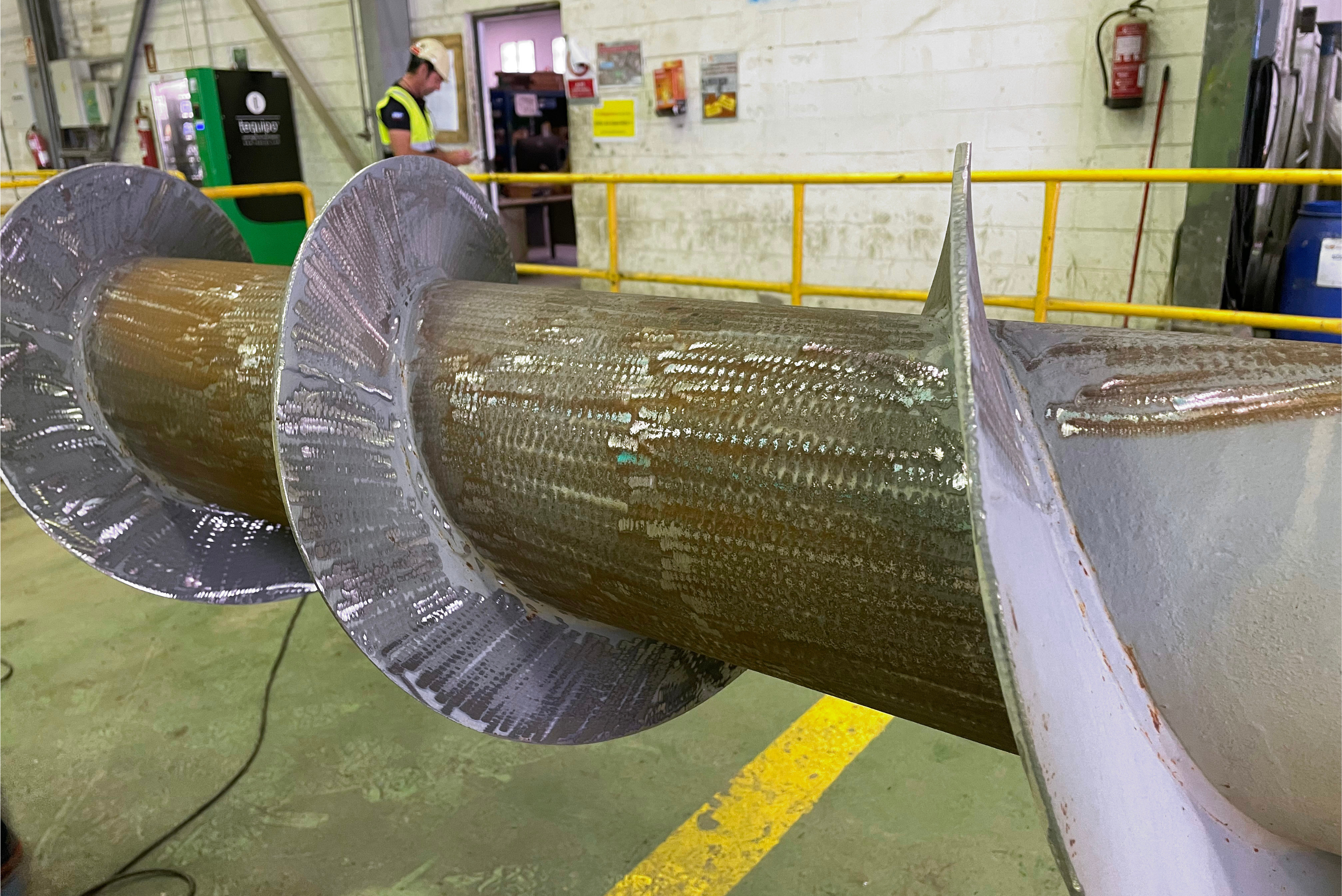 Un grande rotore o coclea industriale con pale elicoidali, parzialmente rivestito con un materiale protettivo grigio, posizionato all'interno di un'officina con una ringhiera gialla sullo sfondo.