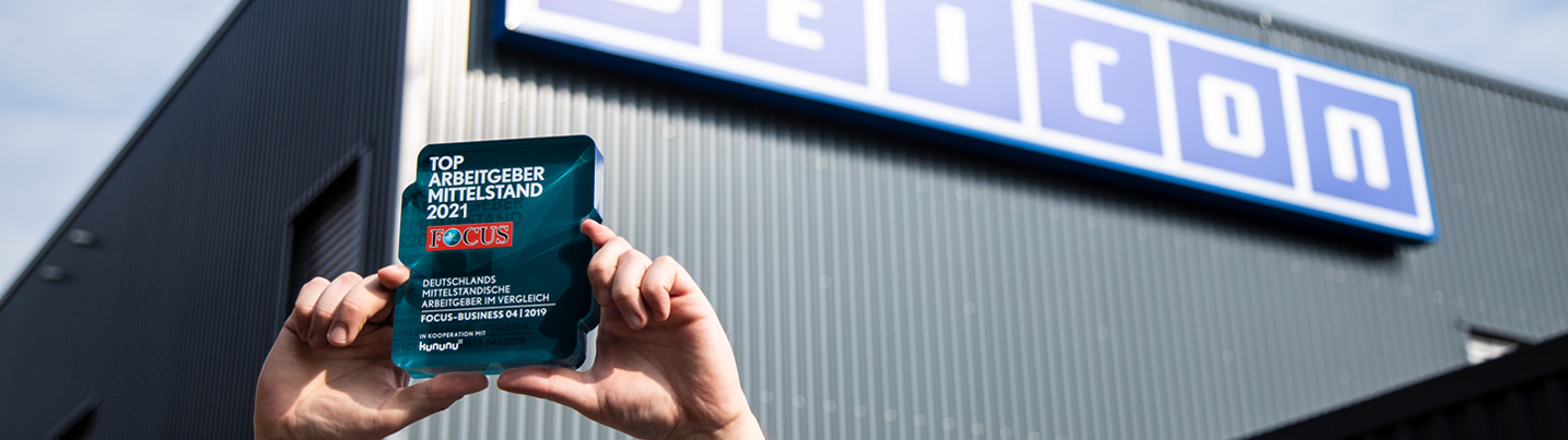 Leitlinien zertifikate Zwei Hände halten eine Trophäe mit "Top Arbeitgeber Mittelstand 2021! hoch, im Hintergrund ist ein Weicon-Gebäude zu sehen