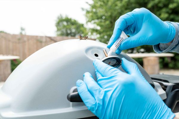 Application de pâte Anti-Seize WEICON sur la tige filetée d'un thermomètre couvercle de barbecue par un opérateur en gants bleus.