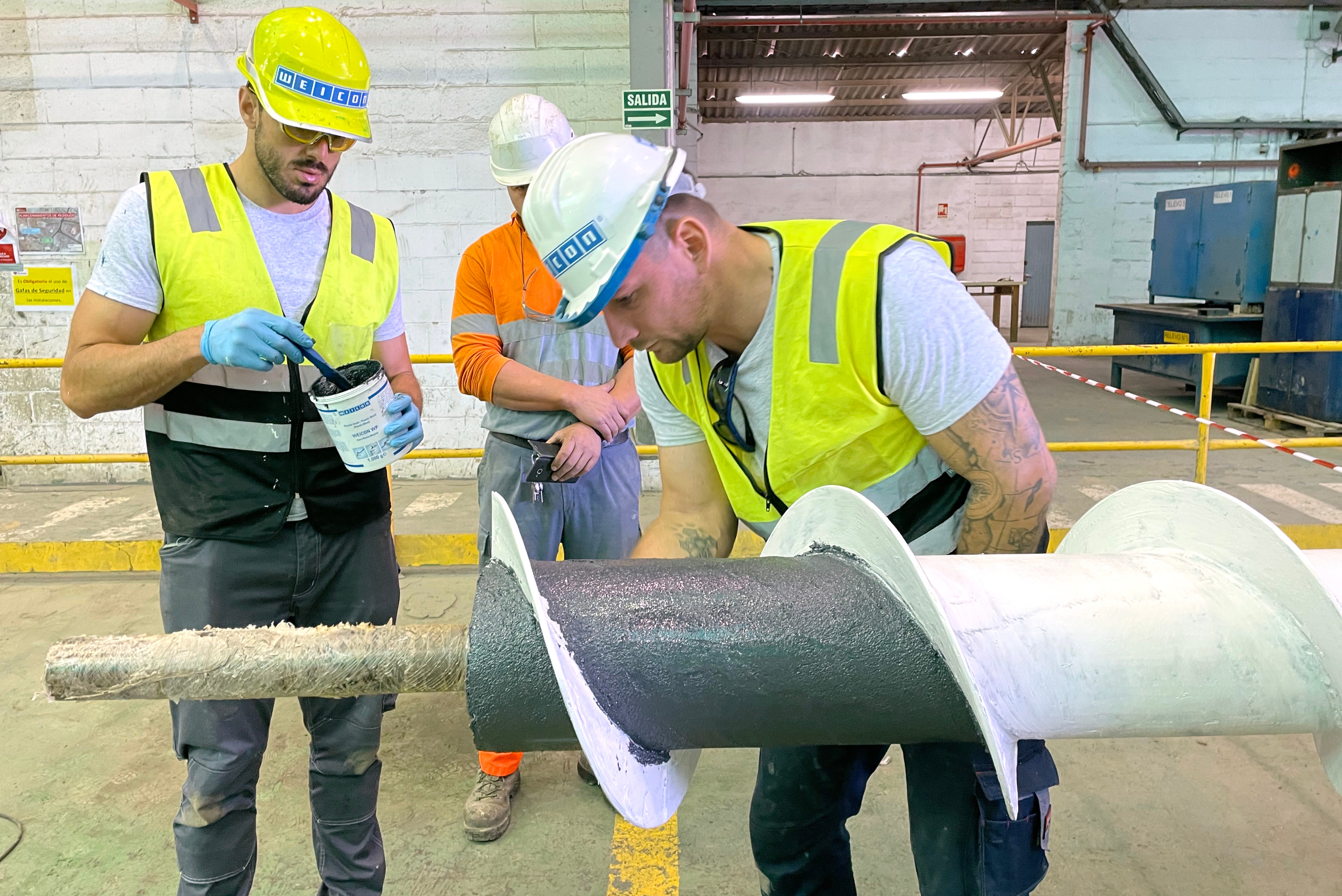 Tre lavoratori in abiti da lavoro, elmetti di protezione e giubbotti ad alta visibilità lavorano su una grande coclea industriale all'interno di un'officina,